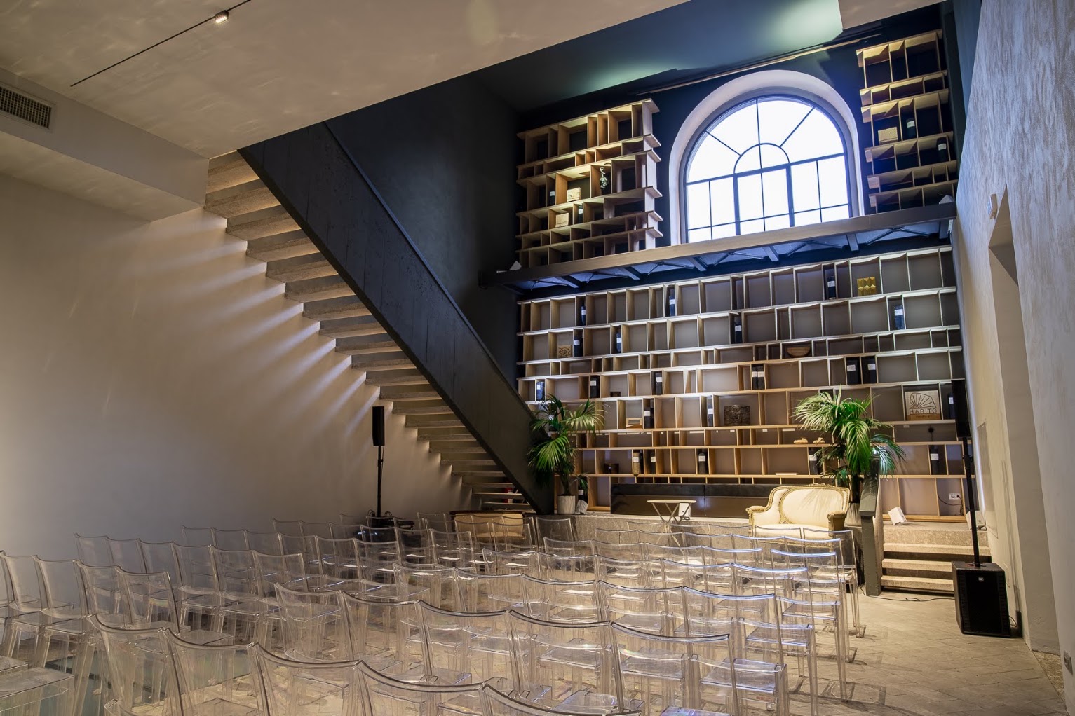 Sala moderna con gradinate di sedie trasparenti, pareti con scaffali e grande finestra ad arco.. location a brera per fashion week, party, formazione, conferenze